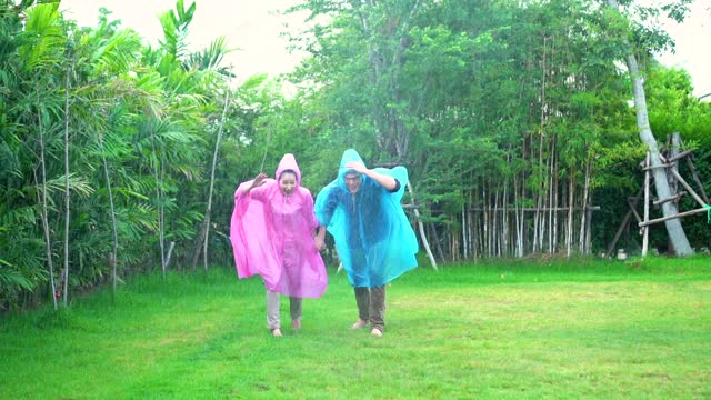 快乐的亚洲夫妇穿着雨衣在户外公园玩雨。视频素材