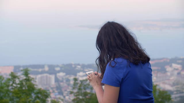 深色头发的女人在城市风景的山顶上使用手机。旅行的女人看智能手机上的山峰，城市和海洋景观视频素材