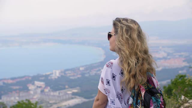 快乐的女游客在山顶欣赏海景。微笑的女人从山顶的观景台欣赏美丽的风景到度假城市。视频素材