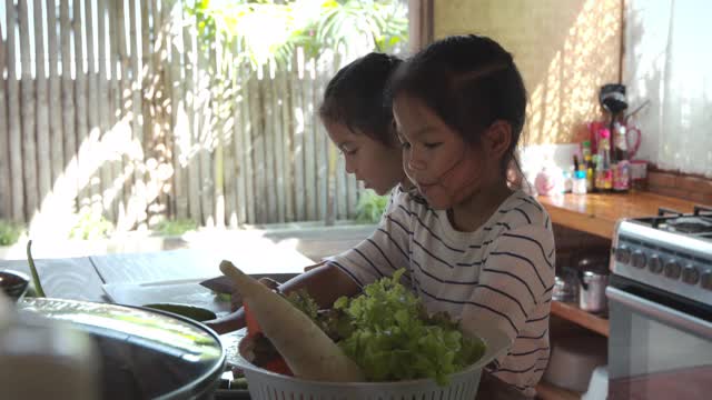 两个可爱的亚洲女孩在厨房里快乐地帮父母布置菜和准备食材，然后一起做家庭晚餐的蔬菜视频素材