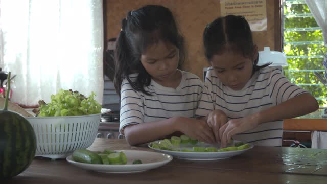 两个可爱的亚洲女孩在厨房里快乐地帮父母布置菜和准备食材，然后一起做家庭晚餐的蔬菜视频素材