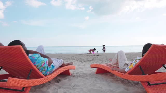 4K混合种族家庭假期快乐。妈妈和爸爸坐在沙滩椅上休息，看着两个女儿在夏天一起坐在沙滩上玩沙子。全家欢度暑假视频下载