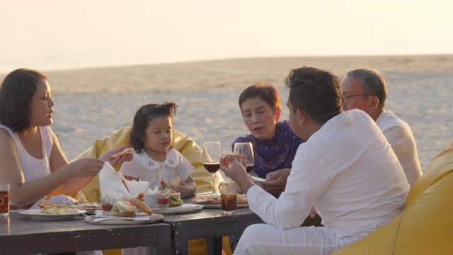 4K多代亚洲家庭在日落时分的海滩上共进晚餐。不同的家庭与孩子女孩，成人和老年夫妇放松和一起享受暑假的乐趣视频下载