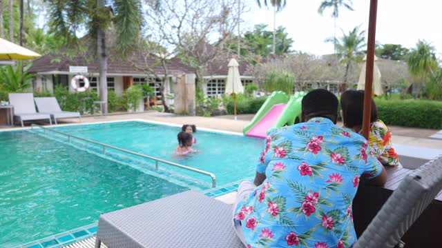 4K混合种族家庭假期快乐。夏天，父母坐在沙滩椅上看着两个可爱的小女孩在酒店的游泳池里一起游泳和玩耍。在夏天的周末，家人放松和玩乐视频下载