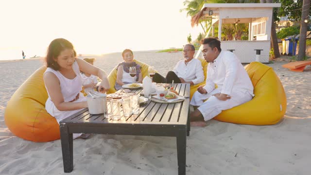 4K多代亚洲家庭在日落时分的海滩上共进晚餐。不同的家庭与孩子女孩，成人和老年夫妇放松和一起享受暑假的乐趣视频下载