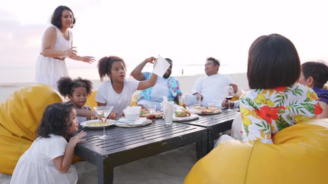 4K一群多民族家庭朋友在日落时分一起在海滩上享用晚餐。不同的大家庭与孩子女孩，成人和老年人夫妇放松和一起玩在暑假假期。视频下载