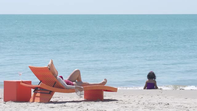 4K亚洲家庭假期快乐。亚洲母亲休息在沙滩椅上看着小女儿在沙滩上玩在夏天的一天。可爱的小女孩放松，和妈妈一起在海滩上玩视频下载