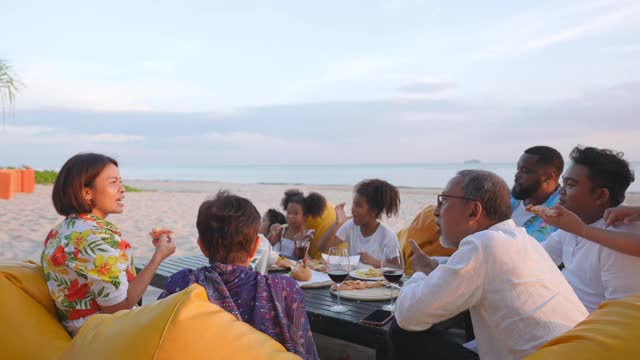 4K一群多民族家庭朋友在日落时分一起在海滩上享用晚餐。不同的大家庭与孩子女孩，成人和老年人夫妇放松和一起玩在暑假假期。视频下载