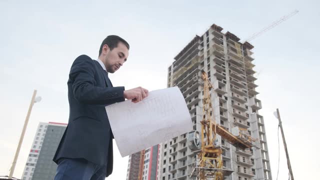 一位年轻的男建筑建筑师正在检查大楼的施工情况。一个工程师看着图纸，背景是街道上的建筑，背景是一座未完工的高层建筑视频素材