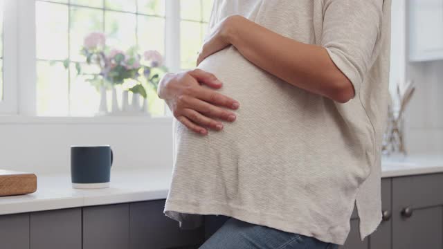 近距离观察孕妇站在厨房窗户旁，用热饮料触摸腹部放松视频素材