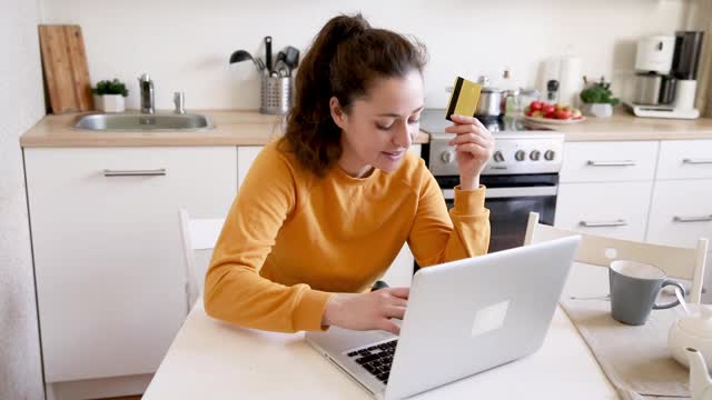 女人在网上购物，用金卡付款。年轻女孩坐在笔记本电脑上上网购买输入信用卡细节厨房室内背景。网上购物电子商务概念视频素材