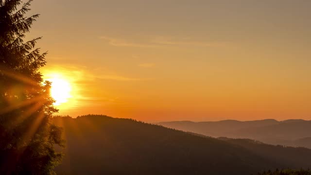 夕阳在橘黄色山丘的地平线上缓缓落下，左侧是一棵针叶树，背景是贝斯基迪(Beskydy)乡村上空的云朵在移动。视频素材