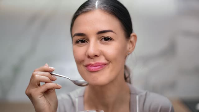 特写微笑的年轻女子在厨房用勺子吃酸奶。用RED摄像机拍摄4K视频素材