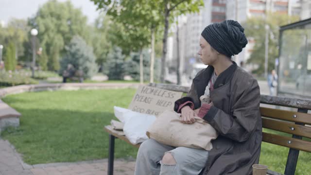 无家可归的女人在肮脏的袋子里寻找食物，坐在长凳上，生存视频素材