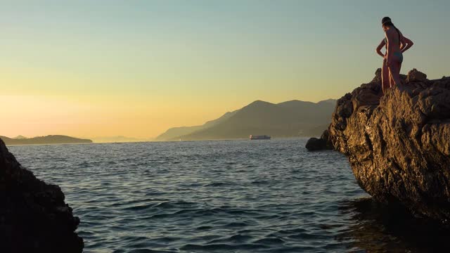 在克罗地亚杜布罗夫尼克附近亚得里亚海海岸的夕阳下，两名少女手拉手从一个高悬崖上跳下视频素材