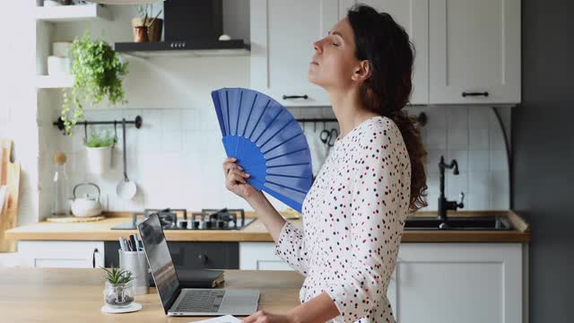 女人站在厨房里，挥动着蓝色的扇子给自己降温视频素材