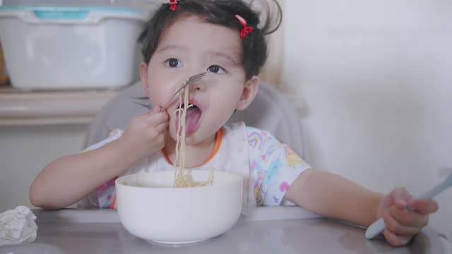 饥饿的亚洲蹒跚学步的孩子坐在椅子上，享受吃面条，而她的家人和她的女儿呆在家里用手握叉为她的午餐脏乱和肮脏。视频素材