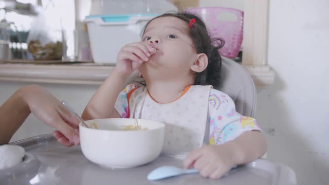 饥饿的亚洲蹒跚学步的孩子坐在椅子上，享受吃面条，而她的家人和她的女儿呆在家里用手握叉为她的午餐脏乱和肮脏。视频素材