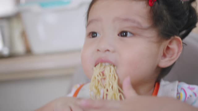 饥饿的亚洲蹒跚学步的孩子坐在椅子上，享受吃面条，而她的家人和她的女儿呆在家里用手握叉为她的午餐脏乱和肮脏。视频素材