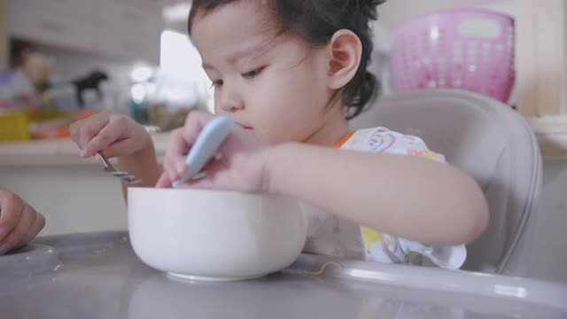 饥饿的亚洲蹒跚学步的孩子坐在椅子上，享受吃面条，而她的家人和她的女儿呆在家里用手握叉为她的午餐脏乱和肮脏。视频素材