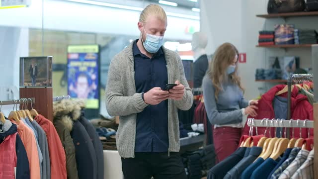 自信的男友戴着口罩在手机上发短信，女友在服装店选购衣服。年轻的白人夫妇在冠状病毒黑色星期五购物。电影4k ProRes总部。视频素材