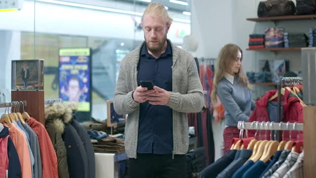 一名留胡子的帅气年轻男子在智能手机上上网，而一名女子在背景中选择着装。一对自信的白人年轻夫妇在购物中心黑色星期五的销售。电影4k ProRes总部。视频素材