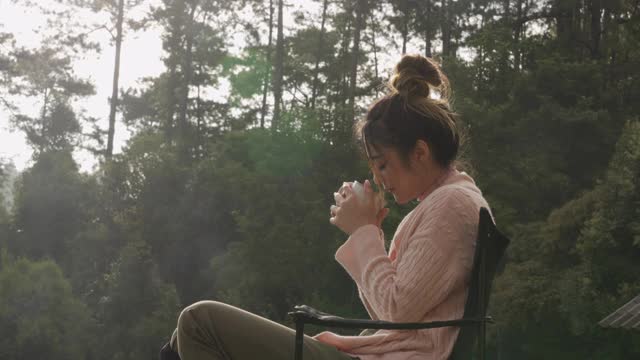4K模糊动作年轻美丽的亚洲女人坐在户外椅子在度假阳台上喝一杯咖啡在秋天的早晨。漂亮女孩放松，享受户外生活方式和假期。视频下载
