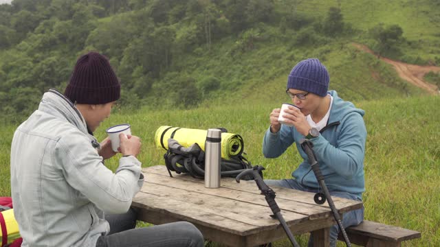 两名亚洲男子喜欢在冬天一起在森林山徒步旅行。快乐的朋友背包客坐在户外的椅子上喝热水。友谊和户外度假概念。视频下载