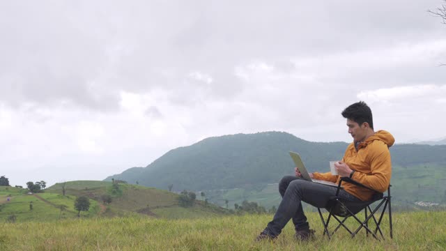 4K亚洲人坐在椅子上用笔记本电脑进行在线工作或视频会议，喝一杯咖啡在山上。帅哥，秋假放松，享受户外生活。视频下载