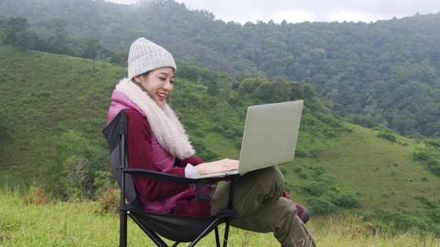年轻美丽的亚洲女子坐在户外椅子上，用笔记本电脑上网进行视频通话或视频博客。美女在寒假放松，享受户外生活。视频下载