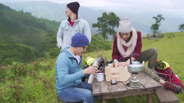 一群亚洲男人和女人背包客喜欢在冬天在森林山一起徒步旅行和露营。快乐的朋友背包客坐在户外的椅子上喝热水。友谊和户外度假概念。视频下载