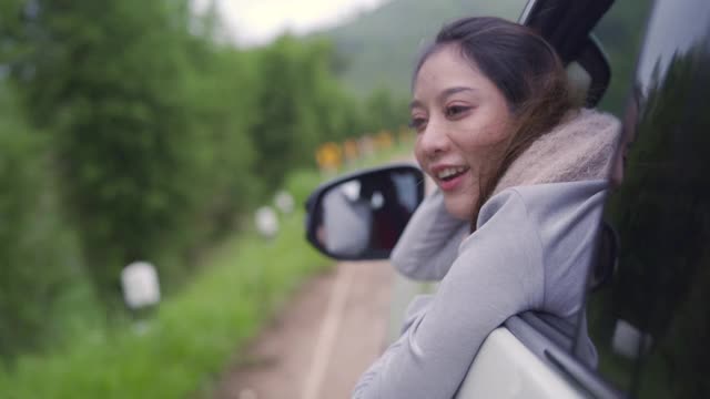 慢动作的快乐年轻美丽的亚洲女人旅行者坐在汽车与拉她的脸和手在雨天的车窗。漂亮女孩喜欢和享受户外生活方式和假期的乐趣。视频下载