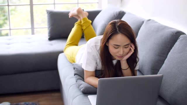 一名女子躺在沙发上用笔记本电脑上网，透过窗户可以看到自然光视频素材