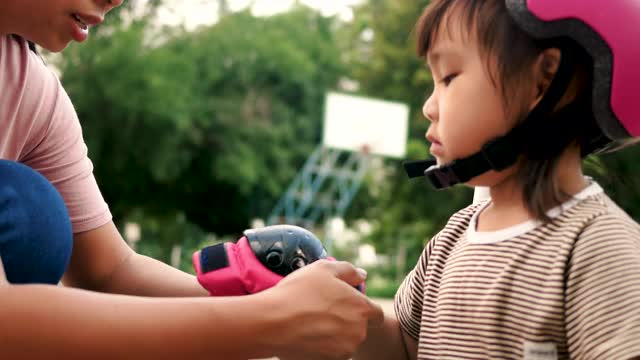 年轻的母亲帮助她的女儿戴上防护头盔和在夏天公园学习骑滑板车之前的肘部。儿童积极的户外运动。视频素材