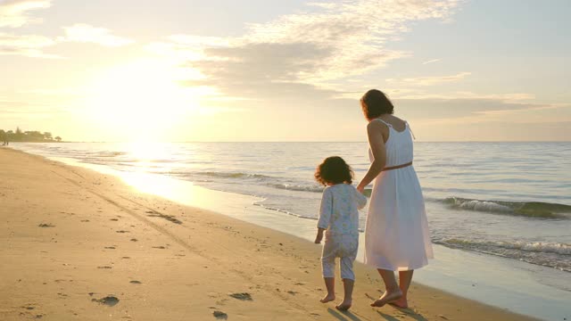 4K后视图的幸福亚洲家庭度假。美丽的母亲穿着白色的连衣裙牵着她的小女儿在夏天的日出一起走在海边。女人与可爱的孩子女孩放松和享受在海滩上。视频下载