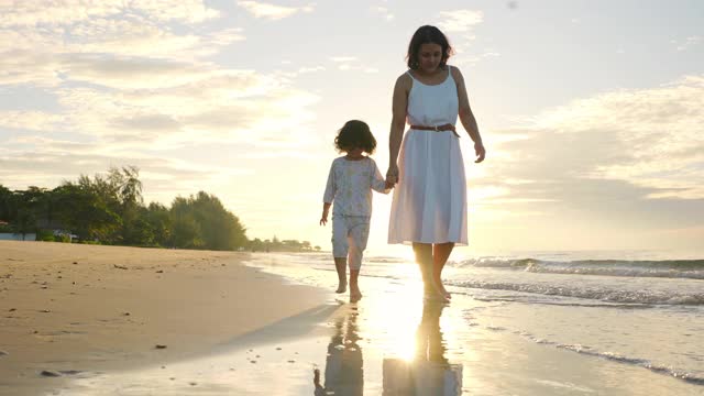 4K快乐亚洲家庭度假。美丽的母亲穿着白色的连衣裙牵着她的小女儿在夏天的日出一起走在海边。女人与可爱的孩子女孩放松和享受在海滩上。视频下载