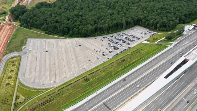 临近多车道高速公路和在建高速公路的半空大停车场。绿植和森林环绕。汽车在路上行驶。空中高角度的视角从空投无人机。摄像机正在向地面移动。视频素材