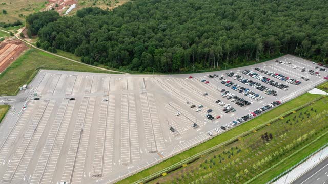 一个大的半空停车场附近的多车道高速公路和建设高速公路。周围是绿色的田野和森林。路上交通方便。从上升的无人机高空角度观看。摄像机在向上移动。视频素材