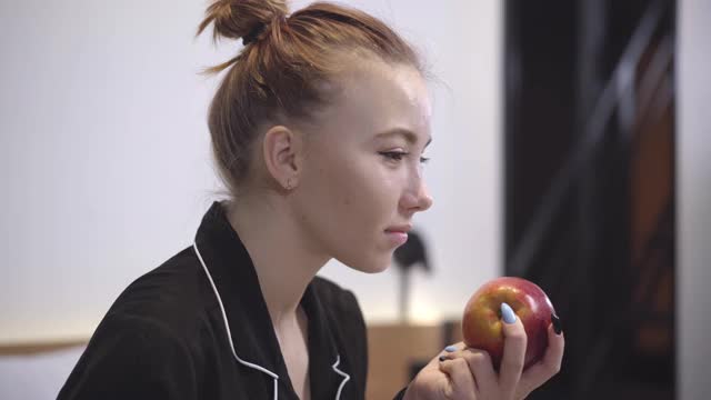 侧面的特写正面美丽的白人女孩吃健康的苹果和喝茶或咖啡在早上。微笑的少年在家厨房吃早餐的肖像。视频素材