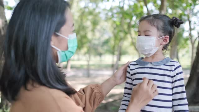 亚洲幼童试图在公共花园摘下卫生口罩。在病毒大流行期间，妈妈帮助她戴上口罩，预防冠状病毒感染。家庭和孩子在户外。健康可爱的理念视频素材