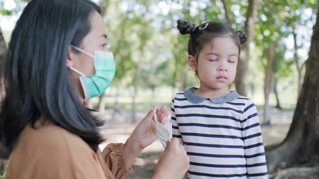 在病毒大流行期间，年轻的亚洲母亲在公园为女儿或孩子佩戴卫生口罩，以预防COVID - 19(冠状病毒)。家庭和孩子在户外。健康可爱的理念视频素材