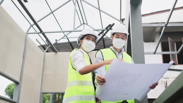 亚洲工人在建筑现场佩戴防护口罩。男和女工程师拿着图纸在施工方讨论项目。视频素材