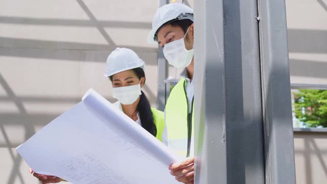 亚洲工人在建筑现场佩戴防护口罩。男和女工程师拿着图纸在施工方讨论项目。视频素材