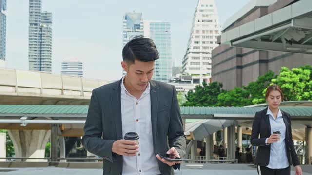 聪明的亚洲年轻商人在城市里使用智能手机。拿着电话的男人微笑着走到外面的办公室。以电信和移动网络为经营理念。视频素材