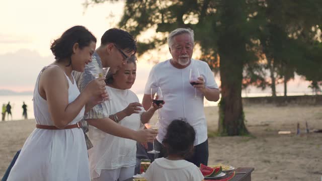 4K亚洲一家三口在夏日日落的沙滩上一起享受晚餐派对和舞蹈。大家庭的小女孩与父母和祖父母放松和有乐趣的假期视频素材