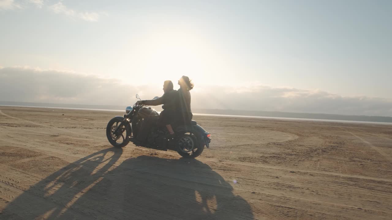 一个男的和一个女的在沙漠里骑摩托车。一对相爱的情侣骑着摩托车在干涸的盐湖上享受日落时分的美好时光。慢动作镜头视频下载