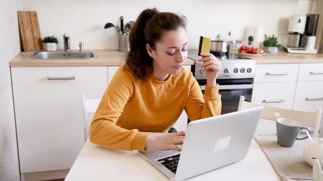 女人在网上购物，用金卡付款。年轻女孩坐在笔记本电脑上上网购买输入信用卡细节厨房室内背景。网上购物电子商务概念视频素材