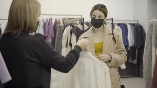 一名感染冠状病毒的自信女子从店员手中拿起装有时髦衣服的衣架，离开更衣室。美丽的年轻白人女性购物肖像。视频素材