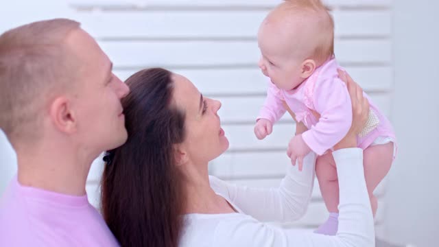 大肖像，一个年轻美丽的家庭，妈妈抱着她的宝贝女儿在她的怀里和亲吻她，爸爸站在她旁边。爸爸妈妈看着宝宝，他们很开心。家庭价值观。4 k, ProRes视频素材