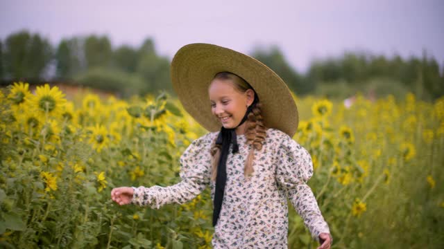 戴着帽子的少女微笑着触摸着夏日漫步在乡村田野上的向日葵。快乐的年轻女孩笑着漫步在黄色的向日葵草地在农村。视频素材